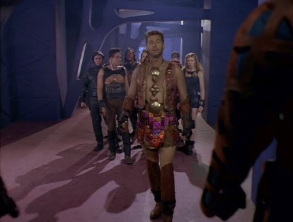 A man wearing a kilt made of little metal tiles that make a gradient of purple to red, a brown vest with matching knee high boots and a medallion that is actually several medallions dangling beneath each other all the way down to his belt.