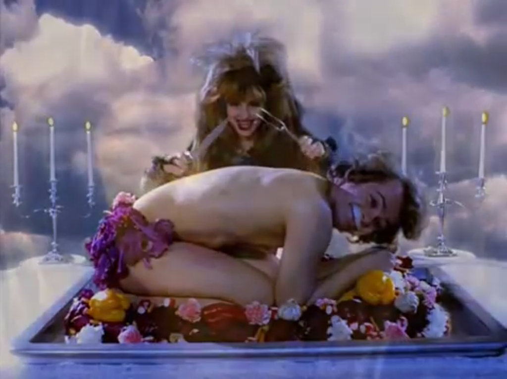 A woman sitting at a table with a large knife and fork about to eat a smiling young lad who's mostly naked and has a milk mustache. he is crouched on a platter piled with vegetables and flowers.