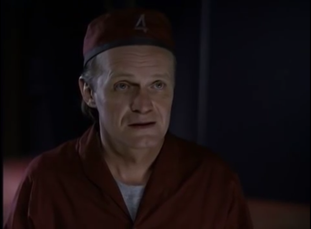 A man with a sad expression on his face. He's wearing a red jumpsuit and a matching red cap with the number 4 on it.