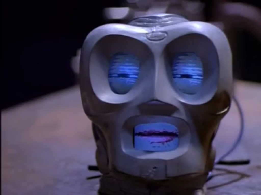 A severed plastic head sitting on a table. The head is a bulbous shape and has big exaggerated circles where the eyes and mouth would be. The eyes and mouth each have a tiny screen in them currently showing squinting eyes and a closed mouth.