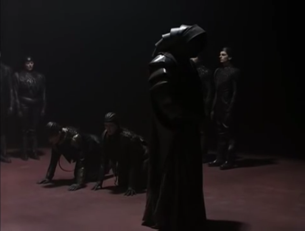 A cloaked figure walks around 2 others that are kneeling on all fours on the floor. Other stand in the background. Everyone is wearing head to to black leather.