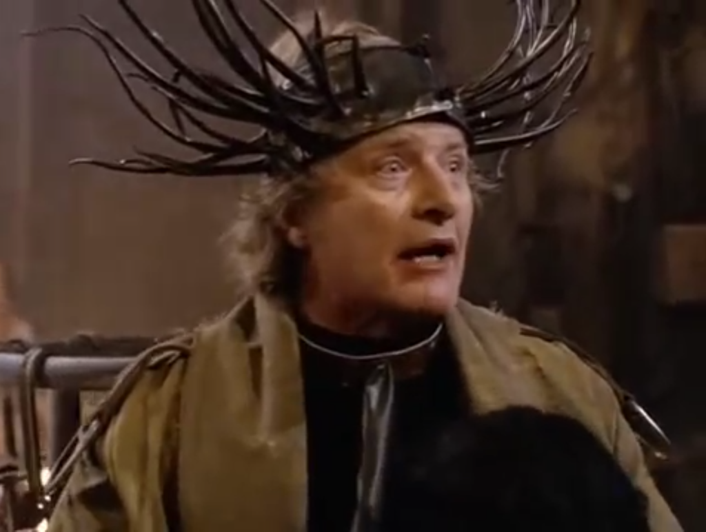 Rutger Hauer wearing a brown coat, metal collar and on his forehead a head piece with numerous spikes coming out of it like antlers or tree branches.