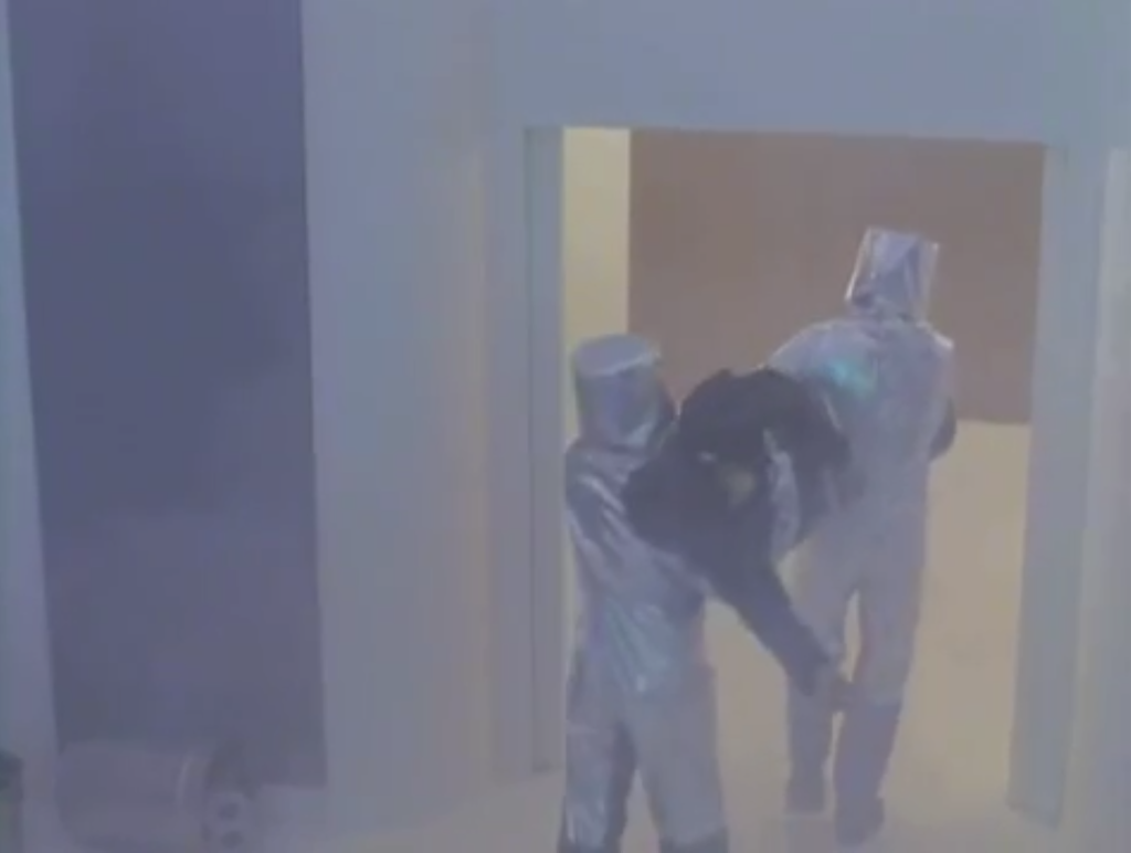 2 men in silver hazmat suits carry off Kai who is frozen and stiff.