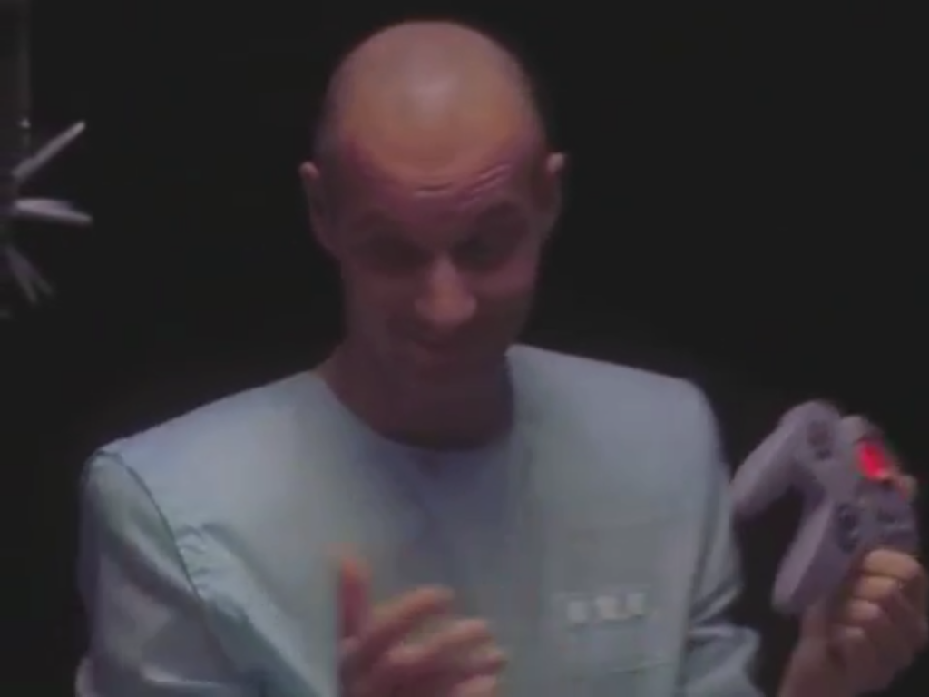 a bald man in scrubs holding a gray Nintendo 64 controller,