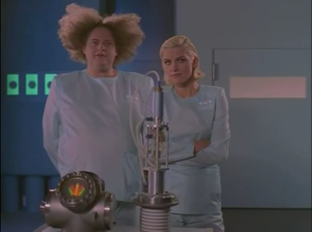 2 doctors in scrubs standing side by side. One is a blonde woman while the other is a man with sandy hair. The man's hair is extremely poofy and curly and is sticking very far out in all directions.