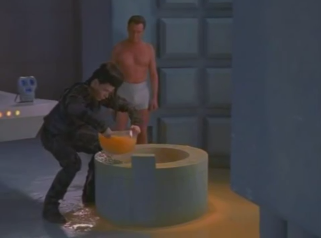 Kai hunched over a small pool in the floor holding a punch bowl of a yellow liquid while Stan and 790 look on.